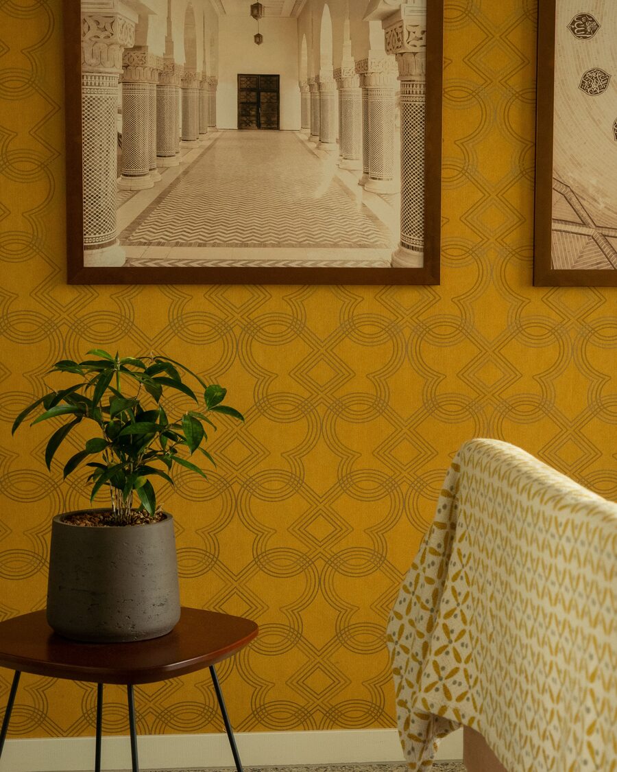 Corner of a sitting room with mustard geometric trellis wallpaper, two framed architectural prints, a potted plant on a side table and the arm of a patterned chair