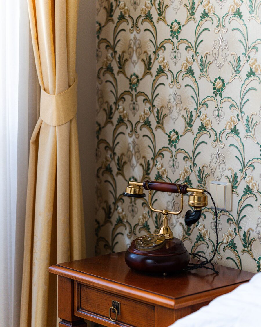 Vintage brass rotary telephone on a cherry side table against a cream damask floral wallpaper beside a gold curtain