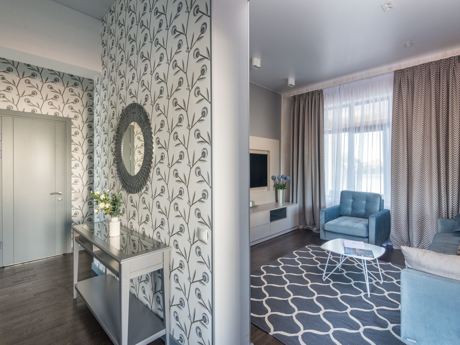 Papered entry hall with a grey botanical vine wallpaper opening onto a living room with a blue armchair and patterned rug