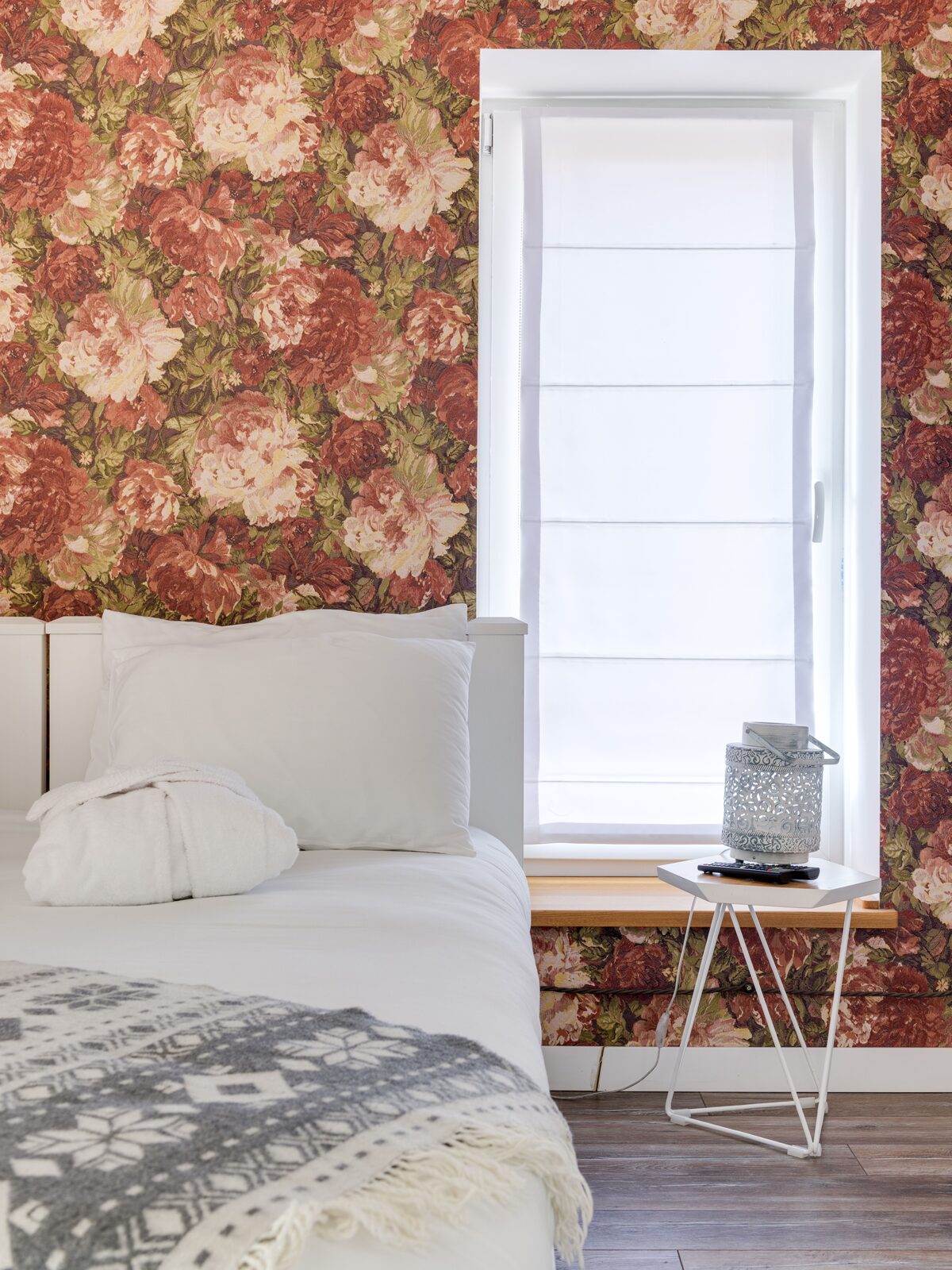 Bright bedroom with a large-scale peony floral wallpaper around a window, white bedding and a knitted throw