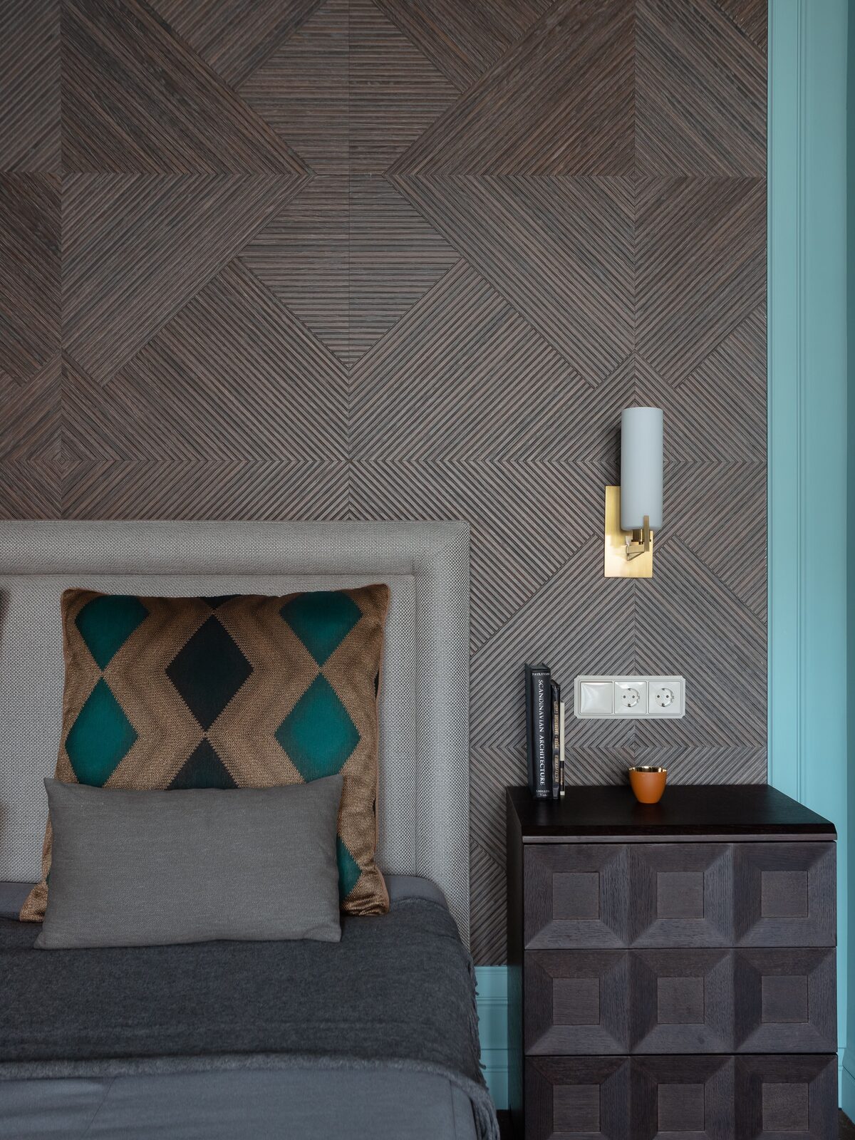 Bedroom feature wall in a geometric wood-grain wallpaper with a brass sconce, upholstered headboard and dark faceted nightstand