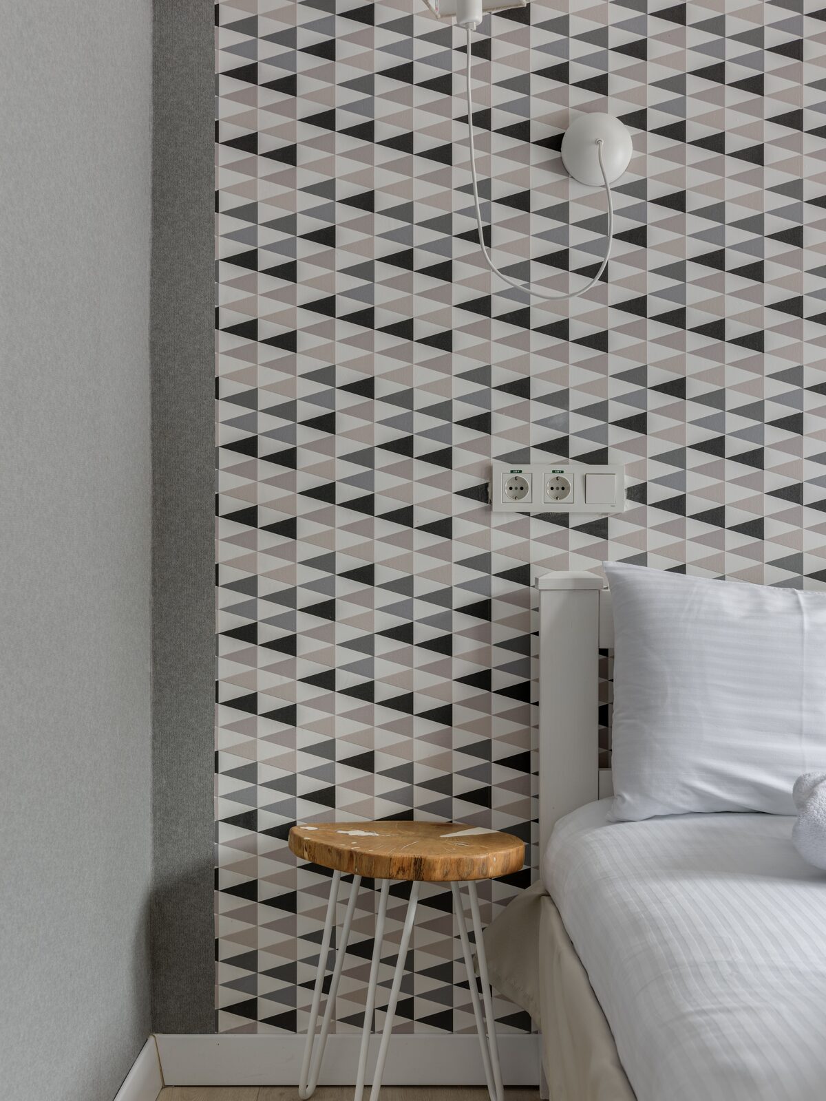 Contemporary bedroom corner with a black, white and grey triangle geometric wallpaper panel, a wooden stool and white bedding