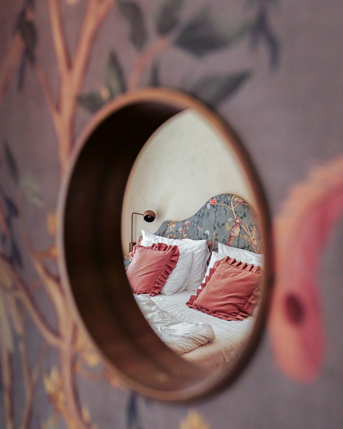 Round brass-rimmed mirror set into a chinoiserie mural wallpaper, reflecting a bed with blush pillows