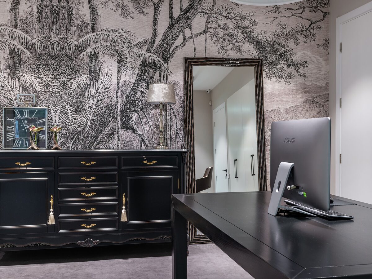 Home office with a black-and-white tropical toile mural wallpaper, black sideboard, framed mirror and a dark desk