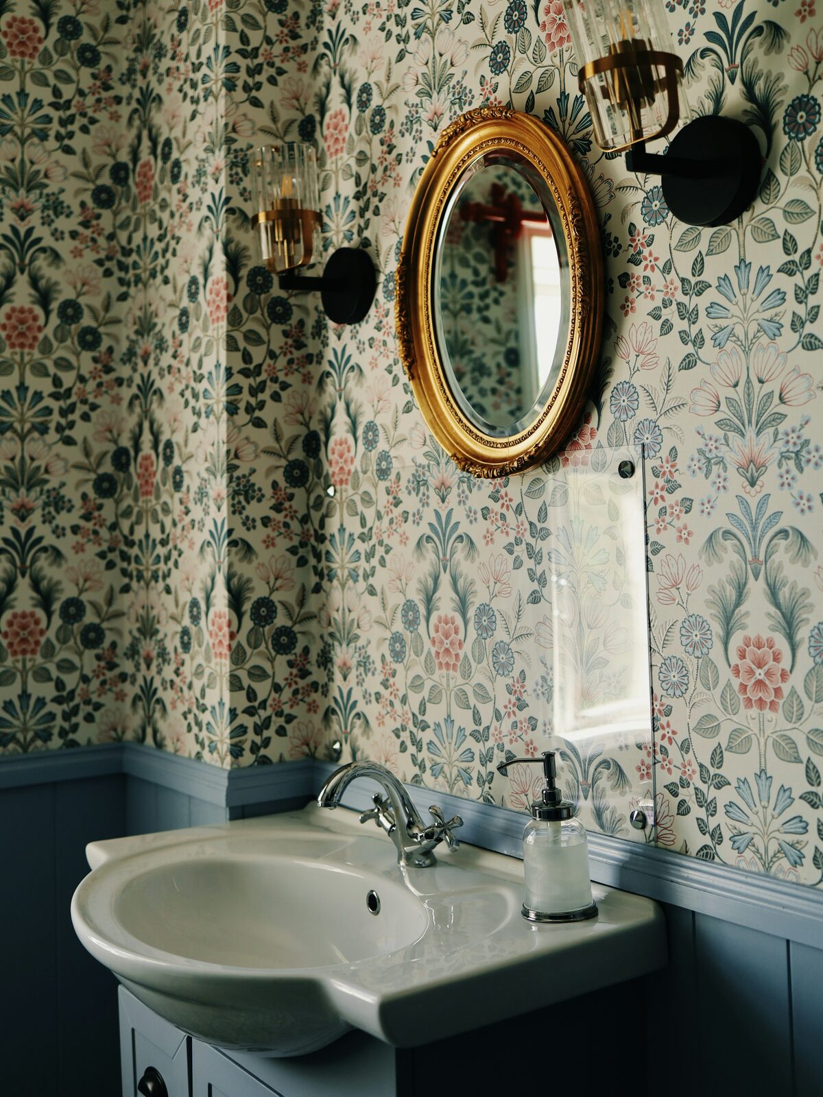 Powder room with a Morris-style floral wallpaper above blue-grey wainscoting, a gilt oval mirror and a pedestal basin