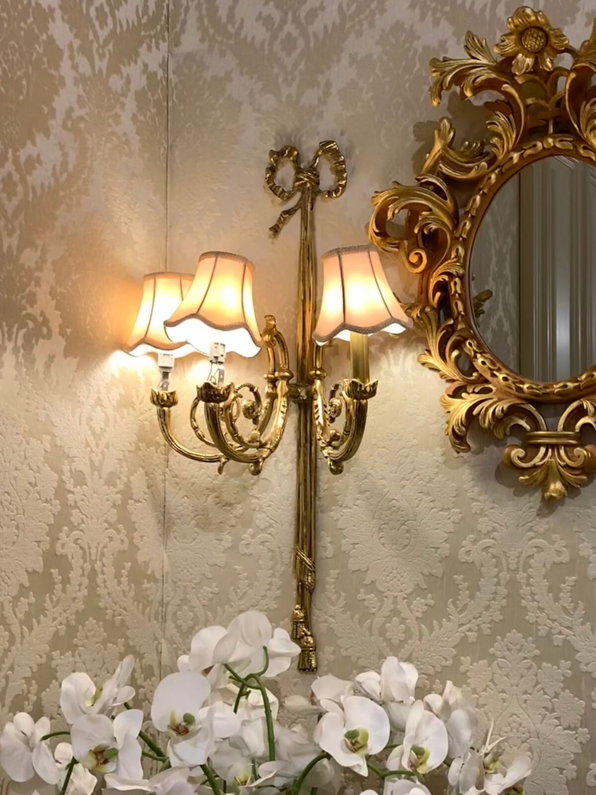 Cream damask wallpaper with a gilt two-arm sconce, an ornate gold mirror and white orchids