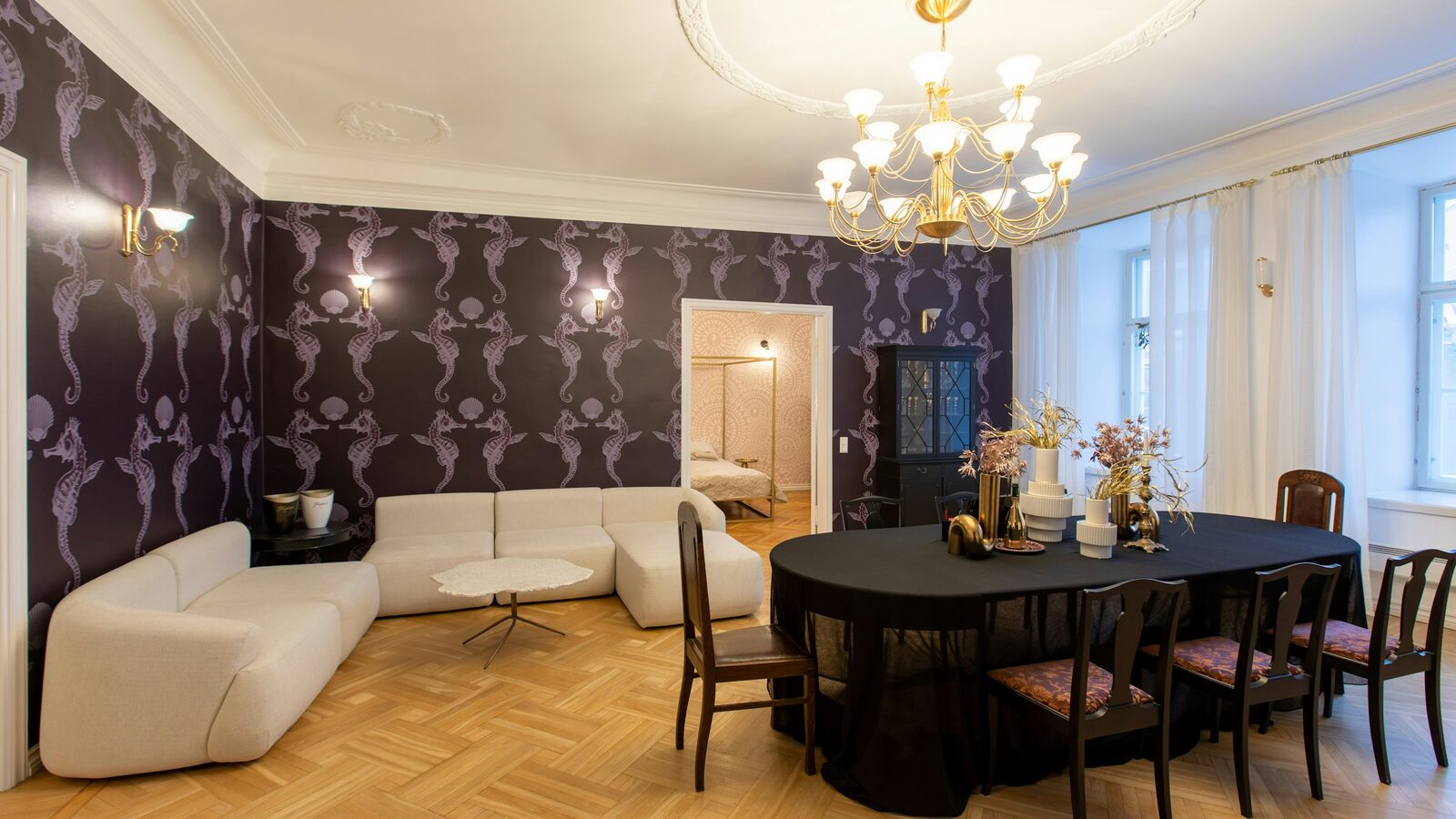 Living and dining room with a deep plum seahorse-pattern wallpaper, white sectional, brass chandelier and herringbone oak floor