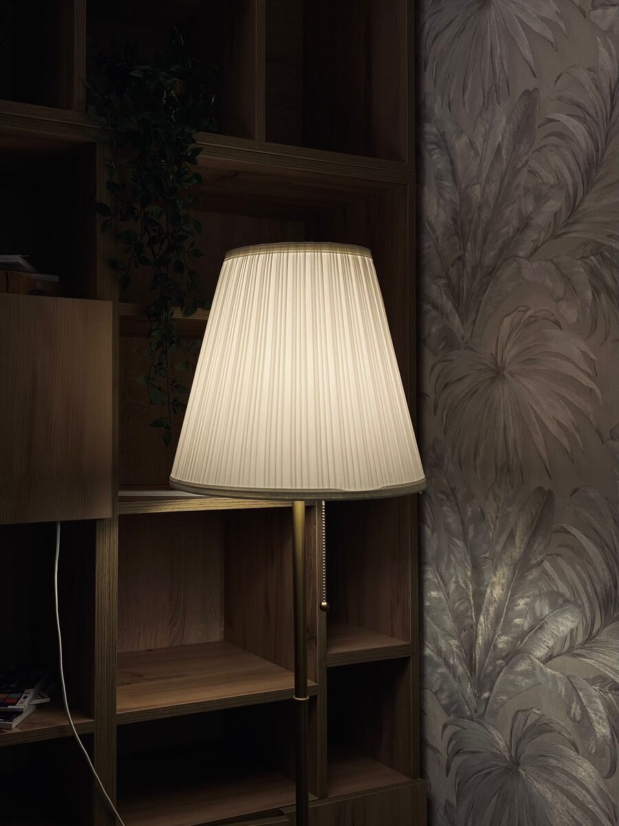 Pleated floor lamp glowing beside dark oak shelving against a taupe palm-leaf wallpaper