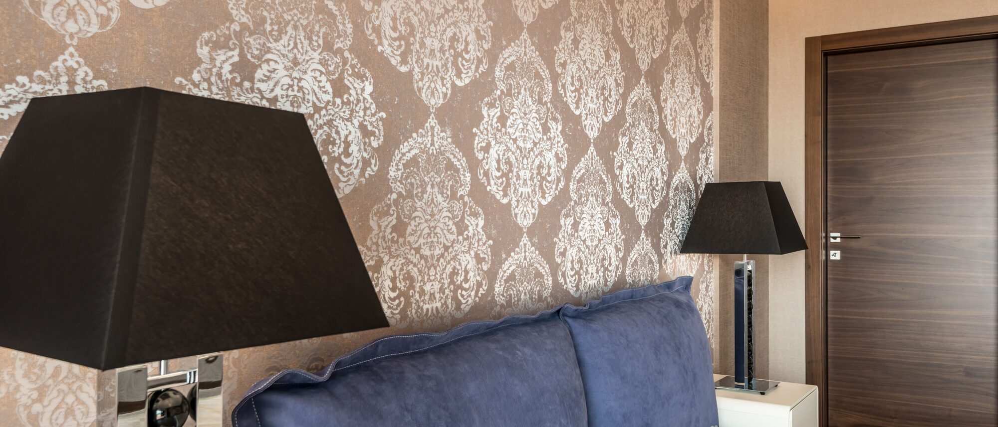 Bedroom feature wall in a bronze metallic damask wallpaper beside a walnut door, with black lamps on either side of a blue velvet headboard